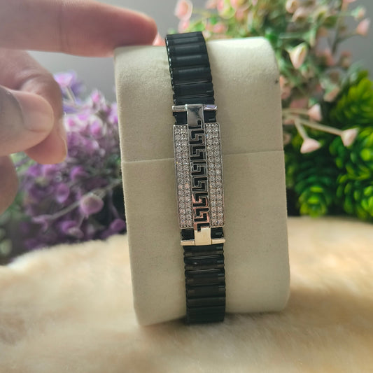 A black and silver bracelet displayed on a cream-colored cylindrical stand, with a hand pointing at it. The background features blurred pink and green flowers.
