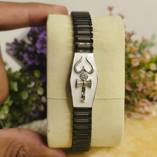 A black and silver coffin-shaped bracelet with an ankh and cross design, held on a cream-colored display block by hands, with blurred flowers in the background.