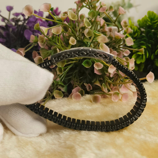 A black braided headband with a silver metal connector, held by a white gloved hand in front of a background of artificial flowers and greenery.