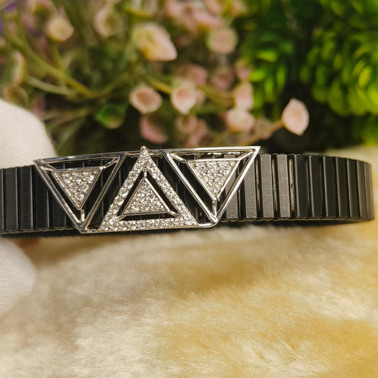 A black ceramic and silver-tone geometric bracelet with triangular crystal-encrusted details, resting on a soft beige faux fur surface with blurred green foliage and pink flowers in the background.