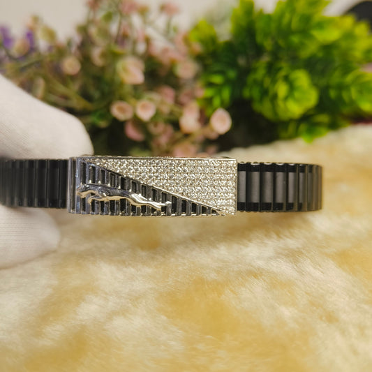 A black and silver cuff bracelet with a central geometric silver plate set with clear crystals and a small silver animal figure, held by a white-gloved hand against a soft beige surface with blurred greenery in the background.