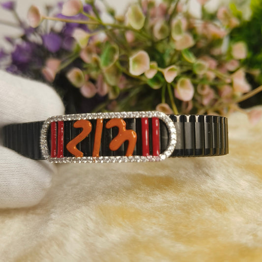 A black metal cuff bracelet with a central rectangular plate featuring red and orange geometric inlay and a border of small clear stones, held by a white gloved hand against a soft beige surface with blurred flowers in the background.