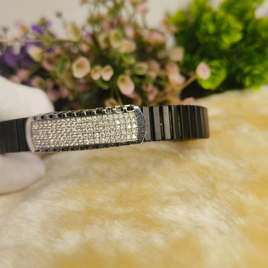 A black and silver striped bangle bracelet with a central rectangular plate paved with clear rhinestones, resting on a soft beige faux fur surface with blurred flowers in the background.