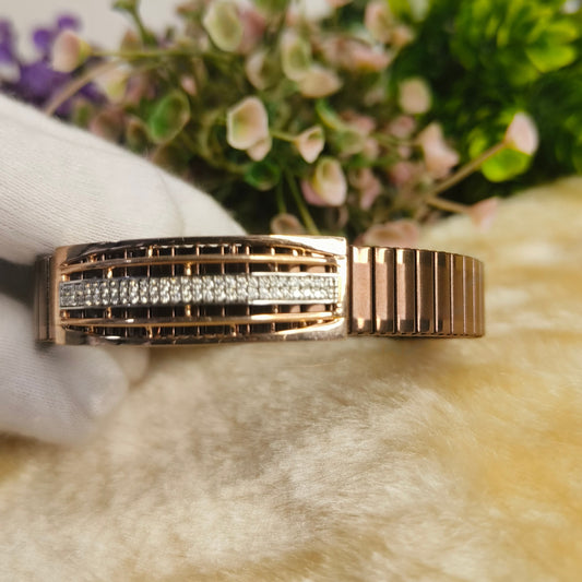 A rose gold cuff bracelet with a central channel of pavé-set clear stones, held by a white-gloved hand against a soft beige faux fur surface with blurred green foliage in the background.
