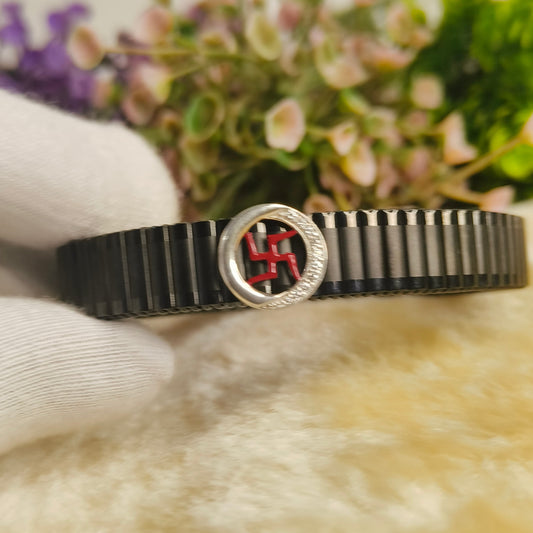 A black segmented bangle bracelet with a circular silver-toned clasp featuring a red central emblem, resting on a white textured surface with a soft beige foreground and blurred flowers in the background.