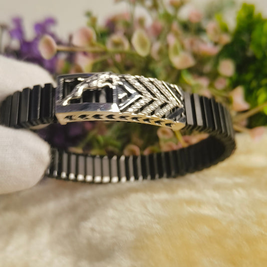 A black and silver geometric bracelet with a horse motif on the clasp, held by a white gloved hand against a soft-focus background of flowers and a beige surface.