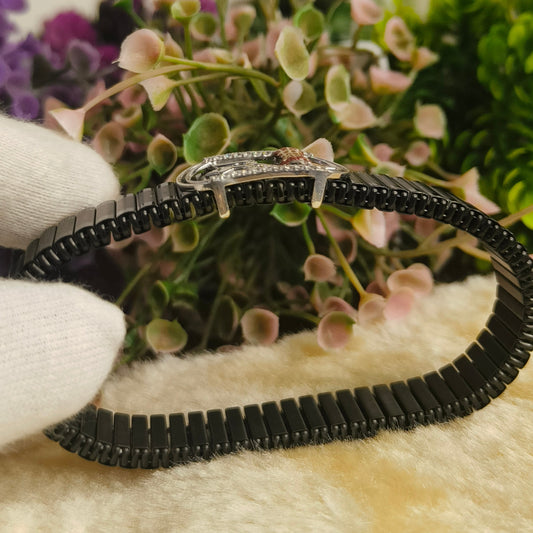 A black beaded bracelet with a silver-toned clasp, held by a white-gloved hand in front of a blurred background of pink flowers and green foliage.