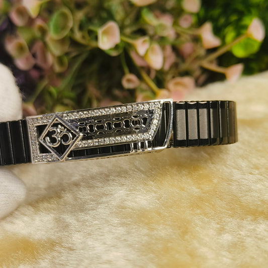 A black and silver Om bracelet with a textured central panel and rhinestone accents, resting on a soft beige surface with a blurred floral background.