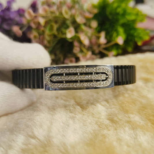A black and silver geometric bangle bracelet with clear crystal-like accents, held by a white-gloved hand against a soft beige faux-fur surface with blurred flowers in the background.
