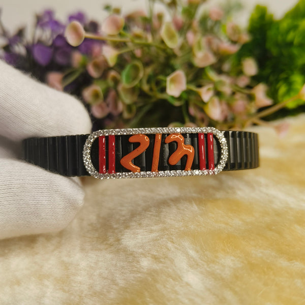 A black bangle bracelet with a rectangular silver-toned centerpiece featuring red and orange decorative elements, held by a white gloved hand against a soft beige surface with blurred flowers in the background.