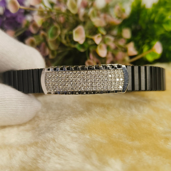A black ceramic or resin bangle bracelet with a rectangular silver-toned bezel set with clear rhinestones, held by a white gloved hand against a soft beige faux fur surface with blurred flowers in the background.