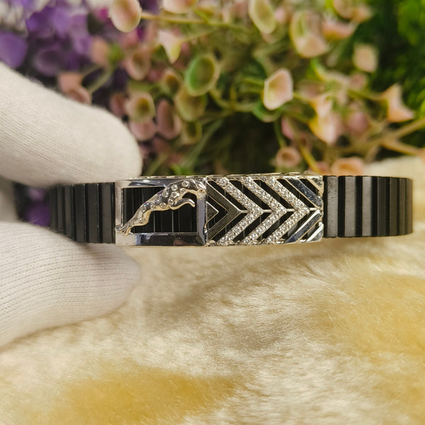 A black and silver striped metal cuff bracelet with a central chevron pattern and horse motif, held by a white-gloved hand against a soft beige surface with blurred flowers in the background.