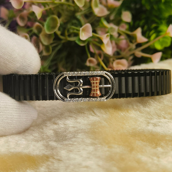A black metal bracelet with a decorative silver-toned central clasp featuring a stylized symbol and a small rose-gold accent, held by a white-gloved hand against a soft beige surface with blurred flowers in the background.