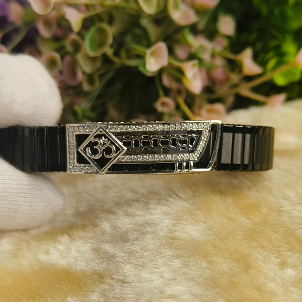 A black ceramic and silver-tone bracelet with a central Om symbol and script engraving, held by a white gloved hand against a soft beige surface with blurred green and pink flowers in the background.